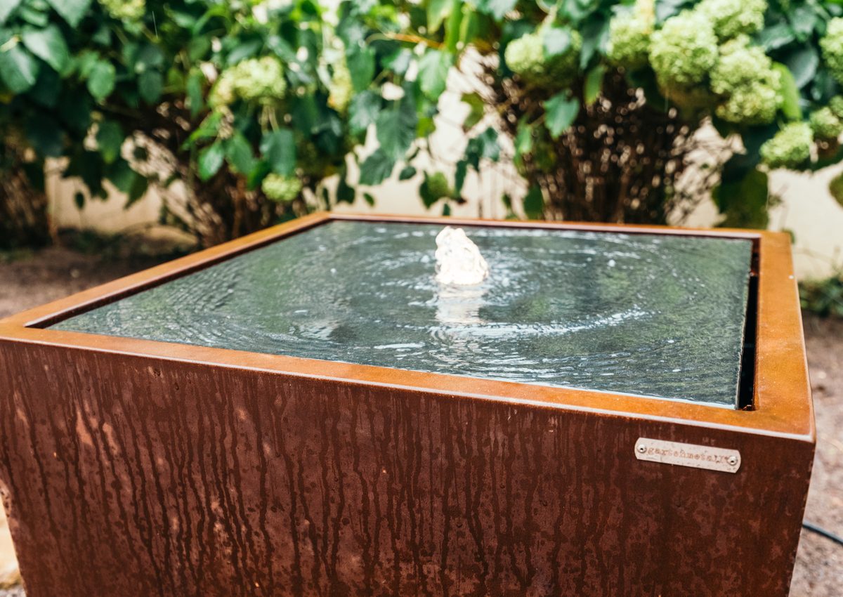 Wasserspiel aus Cortenstahl mit Fontaene vor Hortensien
